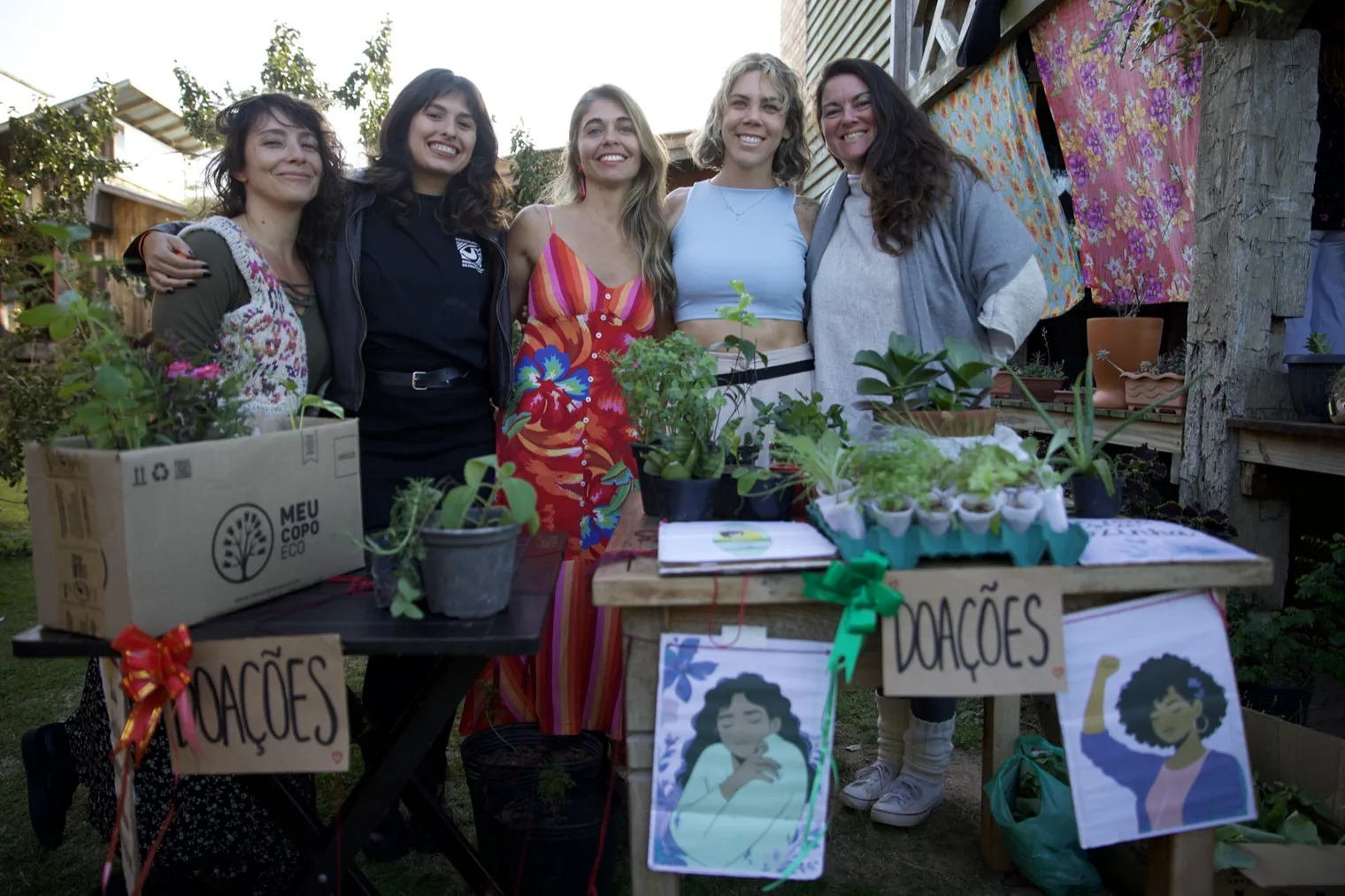Cinco mulheres abraçadas atrás da mesa de doações, cercadas pelas mudas trazidas pelo público, com as placas escritas DOAÇÕES